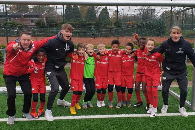 Gruppe junger Fu&szlig;ballspieler in roten Trikots steht l&auml;chelnd vor einem Tor auf einem Fu&szlig;ballplatz.
