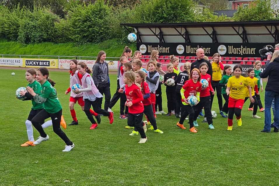 Gruppierung von Kindern in Sportkleidung, die enthusiastisch auf einem Fußballplatz mit Bällen in unterschiedlichen Positionen laufen.
