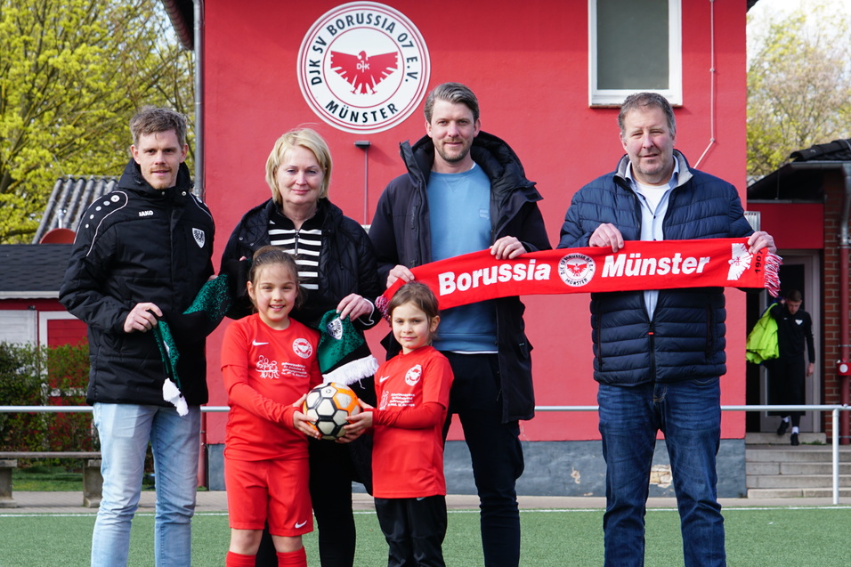 F&uuml;nf Personen stehen auf einem Fu&szlig;ballplatz, halten einen roten Schal mit &bdquo;Borussia M&uuml;nster&ldquo; und einen Fu&szlig;ball.