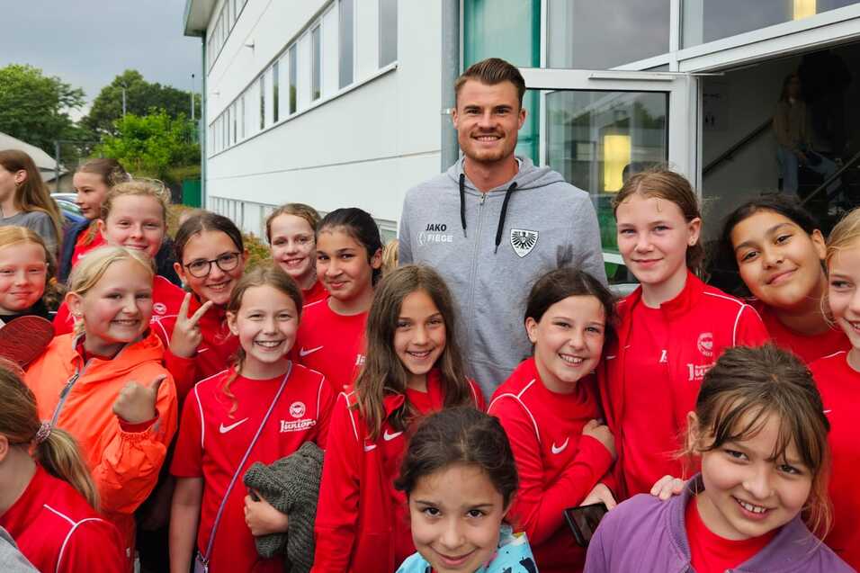 Gruppe von Mädchen in roten Sportjacken posiert mit einem lächelnden Trainer vor einem Veranstaltungsgebäude.