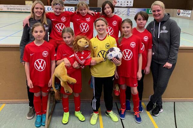 M&auml;dchenfu&szlig;ballmannschaft in roten Trikots mit einer Puppe und einem Ball, umrahmt von Trainerinnen in einer Sporthalle.