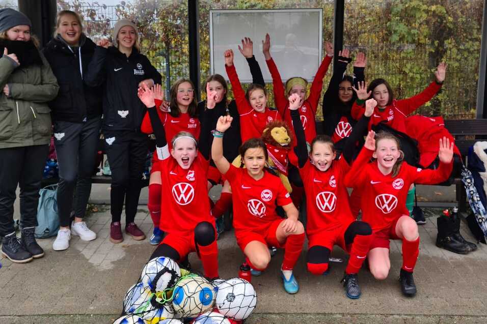 Mannschaft von jungen Fußballspielern in roten Trikots, die fröhlich auf einer Bank stehen, umgeben von Bällen und Betreuern.