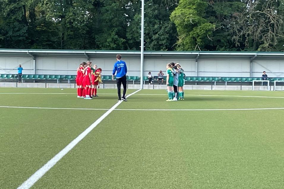 Zwei Fußballmannschaften in Rot und Grün stehen auf einem Kunstrasenplatz im Kreis, eingeladet von einem Trainer.