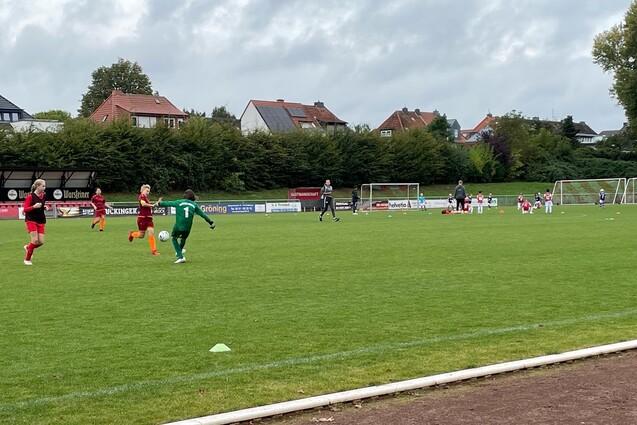 Ein Fu&szlig;ballspiel mit Spielern in roten und gr&uuml;nen Trikots auf einem Rasenplatz, im Hintergrund Tore und Zuschauer.