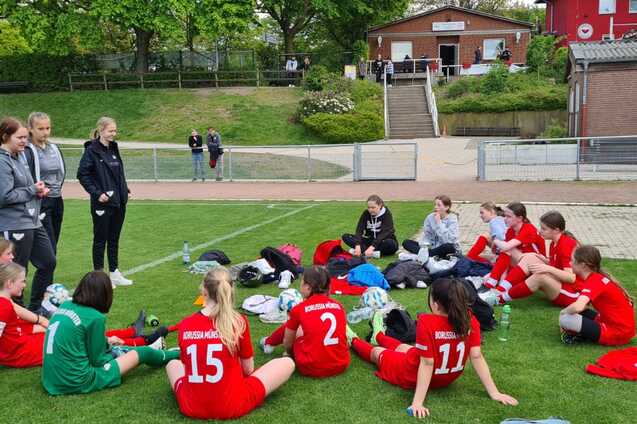 Gruppe von M&auml;dchen in roten Trikots sitzt auf einem Fu&szlig;ballfeld und unterh&auml;lt sich in Teamformation.