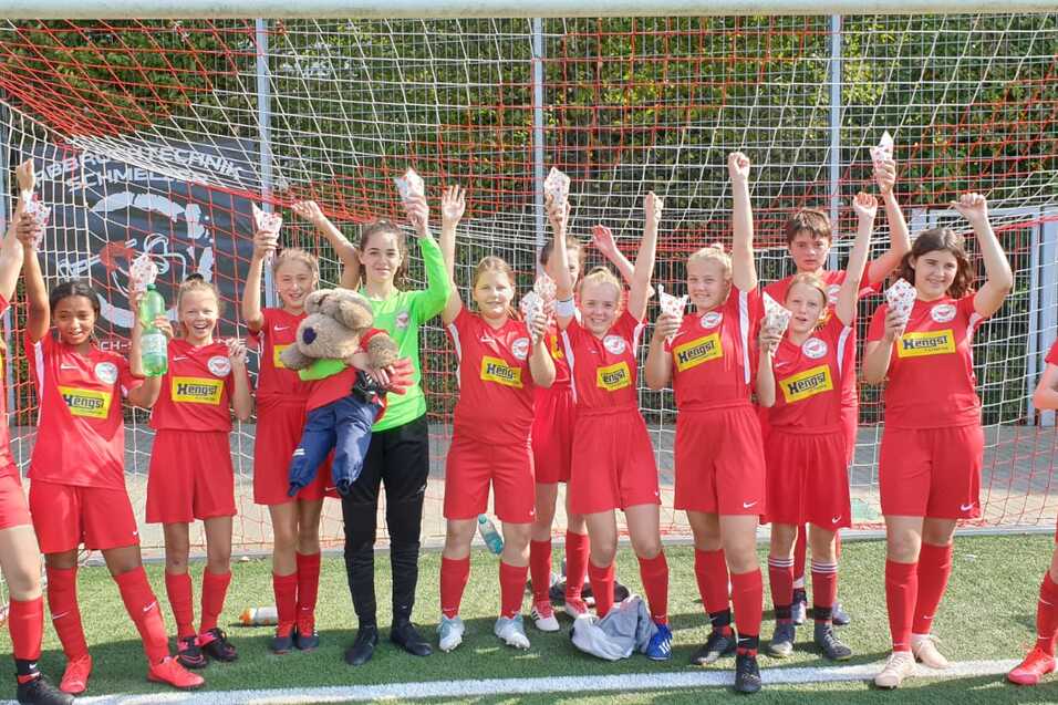 Mädchenfußballmannschaft in roten Trikots feiert vor einem Fußballtor mit Händen in der Luft.
