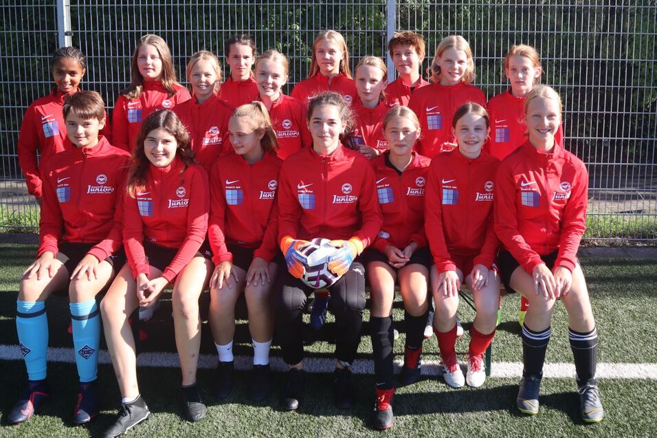 Gruppenfoto eines Fu&szlig;ballteams in roten Trikots auf einem Kunstrasenplatz, mit einer Torfrau und einem Ball im Vordergrund.