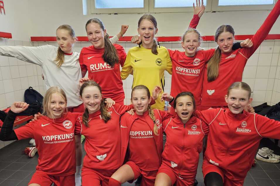 Mädchenfußballmannschaft in roter Sportkleidung posiert fröhlich im Umkleideraum.