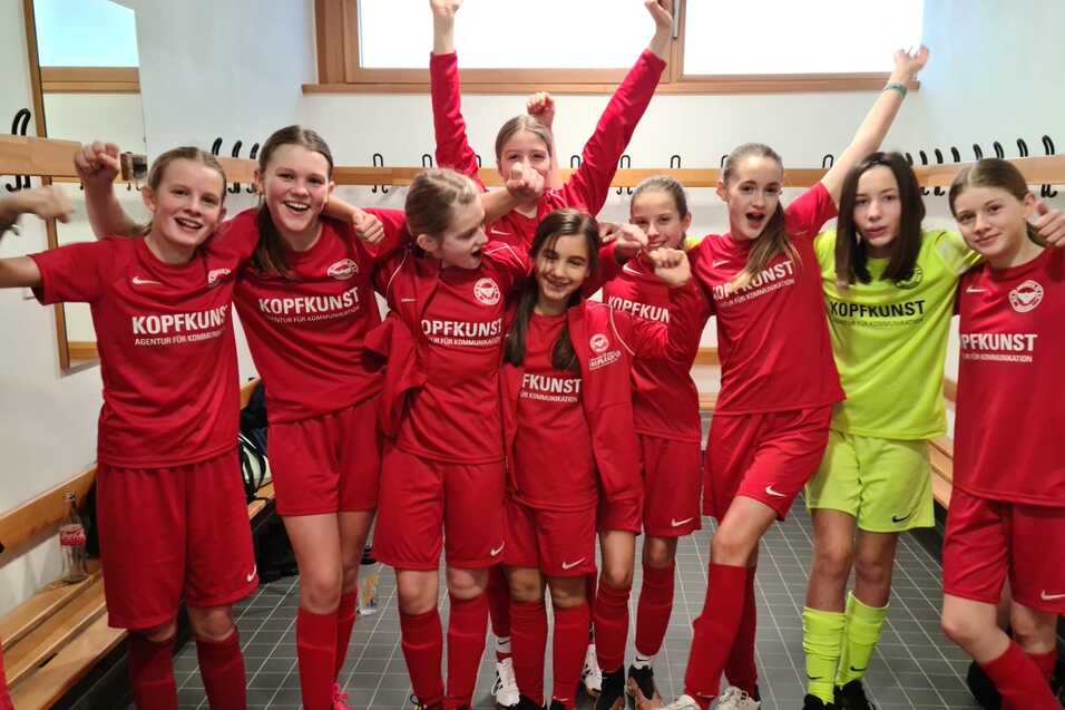 Mädchenmannschaft in roten Fußballtrikots jubelt in Umkleidekabine, einige halten Arme hoch, mit einem Torwart in Gelb.