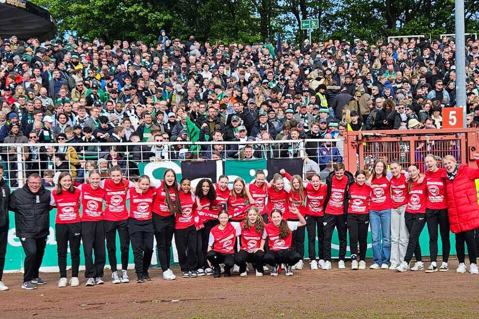 Gruppe von Sportlerinnen in roten Shirts posiert vor jubelndem Publikum im Stadion.
