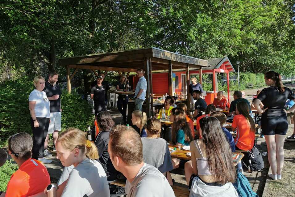 Gruppenszene mit Sportlern, die an Tischen sitzen und essen, umgeben von Gr&uuml;n und einem gem&uuml;tlichen Kiosk.