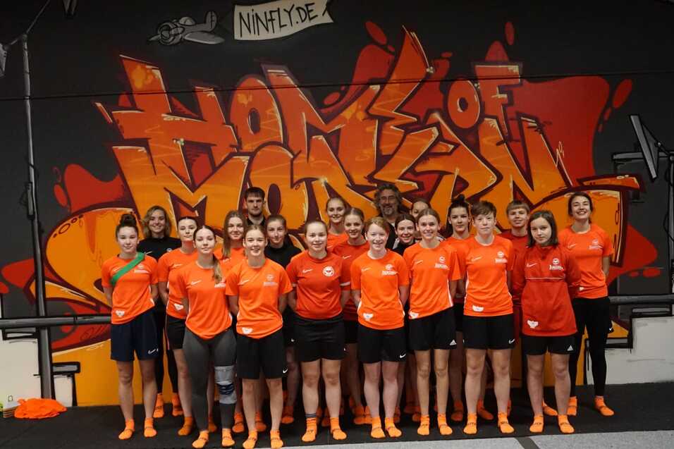 Gruppe von 15 Sportlerinnen in orangefarbenen T-Shirts vor einer graffitiartigen Wand in einer Sporthalle.
