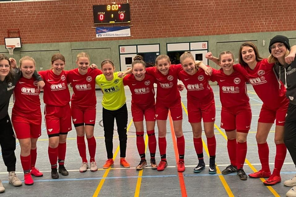 Eine Gruppe von elf Fu&szlig;ballspielerinnen in roten Trikots posiert l&auml;chelnd auf einem Hallenfu&szlig;ballfeld.