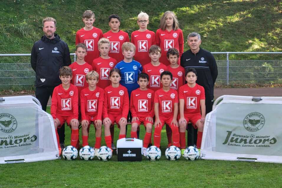 Mannschaftsbild einer Jugendfußballmannschaft in roten Trikots mit Trainern, Bällen und Werbebannern auf dem Rasen.