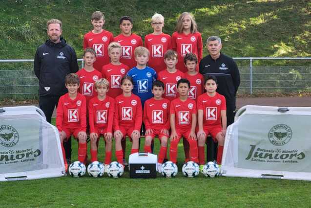 Mannschaftsbild einer Jugendfu&szlig;ballmannschaft in roten Trikots mit Trainern, B&auml;llen und Werbebannern auf dem Rasen.