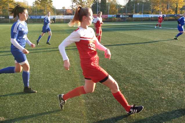Eine Fu&szlig;ballspielerin in roter Uniform f&uuml;hrt mit einem Ball einen Kick auf einem Kunstrasenplatz aus, Spielerinnen im Hintergrund.