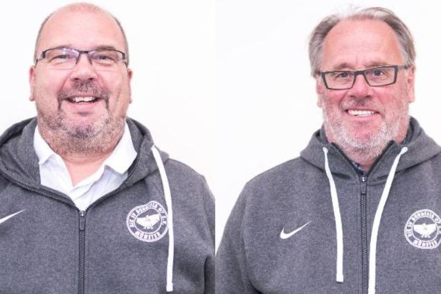 Zwei M&auml;nner in grauen Kapuzenpullis stehen vor einem wei&szlig;en Hintergrund und l&auml;cheln freundlich in die Kamera.