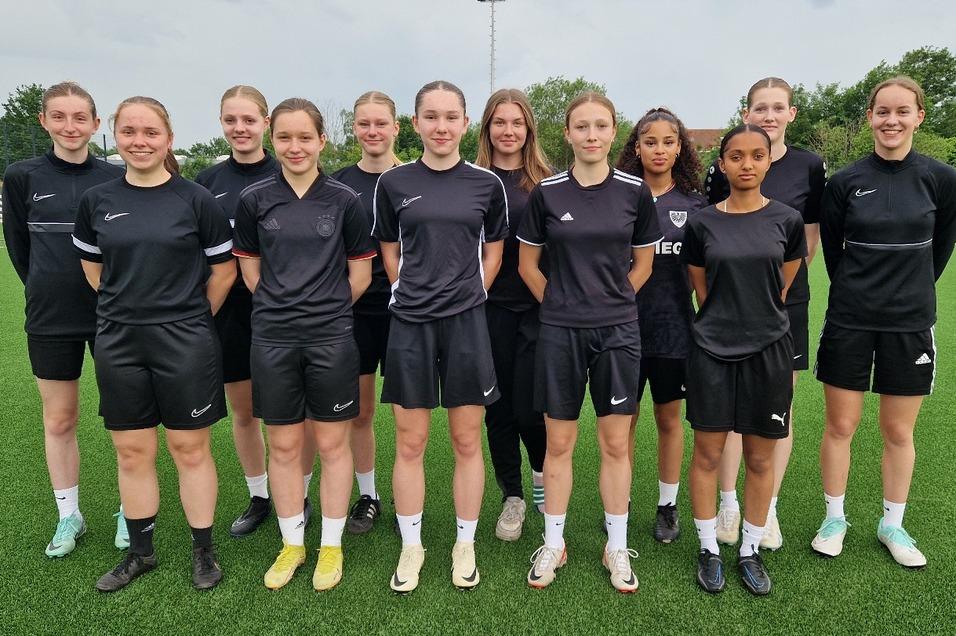 Gruppe von zwölf Frauen in schwarzen Sporttrikots steht auf einem Fußballfeld mit Kunstrasen.