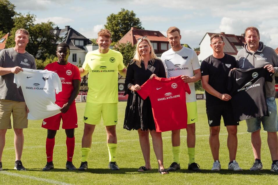 Sechs Personen auf einem Fußballfeld halten Trikots in verschiedenen Farben, im Hintergrund ist eine Wohngegend sichtbar.