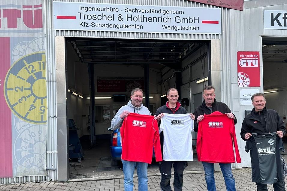 Vier Männer stehen vor einem GTÜ-Gebäude und halten T-Shirts in den Farben Rot und Weiß sowie eine schwarze Tasche.
