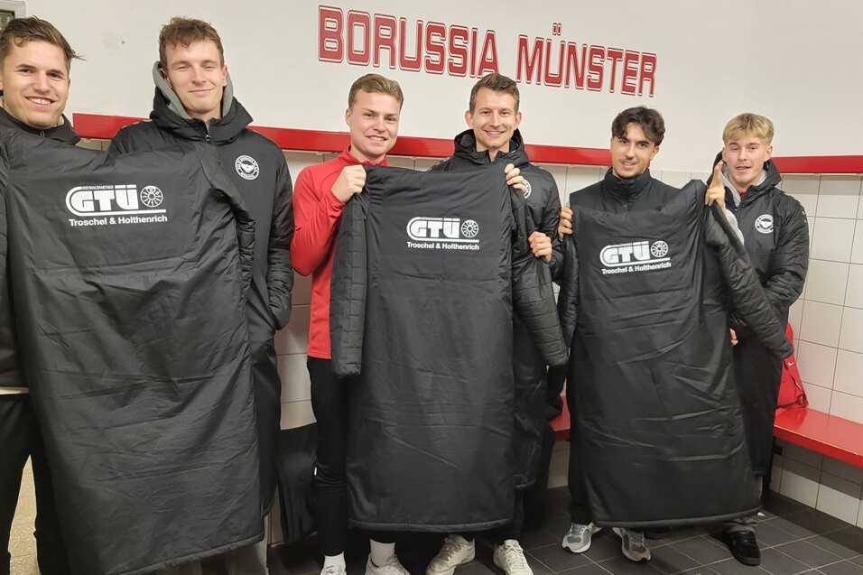 Sechs Sportler in Umkleidung halten schwarze Trikots mit Logo vor dem Schriftzug "BORUSSIA MÜNSTER" in einem Kabinensaal.