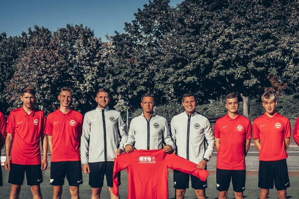 Teamfoto von acht Fußballspielern in roten und grauen Trikots, auf einem Sportplatz mit Bäumen im Hintergrund.