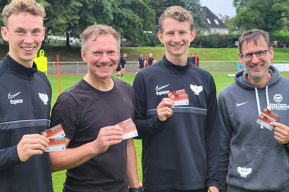 Vier Männer halten stolz ihre Mitgliedskarten vor einem Fußballfeld, umgeben von Bäumen und einem Sportplatz.