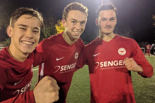 Drei junge Fu&szlig;ballspieler in roten Trikots posieren l&auml;chelnd auf einem beleuchteten Platz bei Nacht.