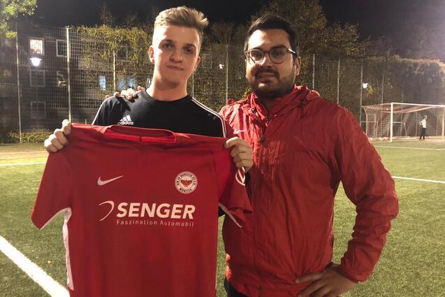 Zwei M&auml;nner stehen auf einem Fu&szlig;ballplatz bei Nacht und halten ein rotes Trikot mit Sponsorenlogo hoch.