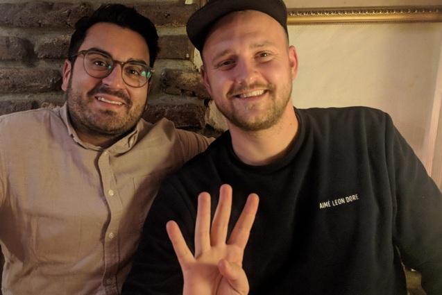 Zwei M&auml;nner sitzen an einem Tisch, einer zeigt mit der Hand die Zahl vier, beide l&auml;cheln in die Kamera.