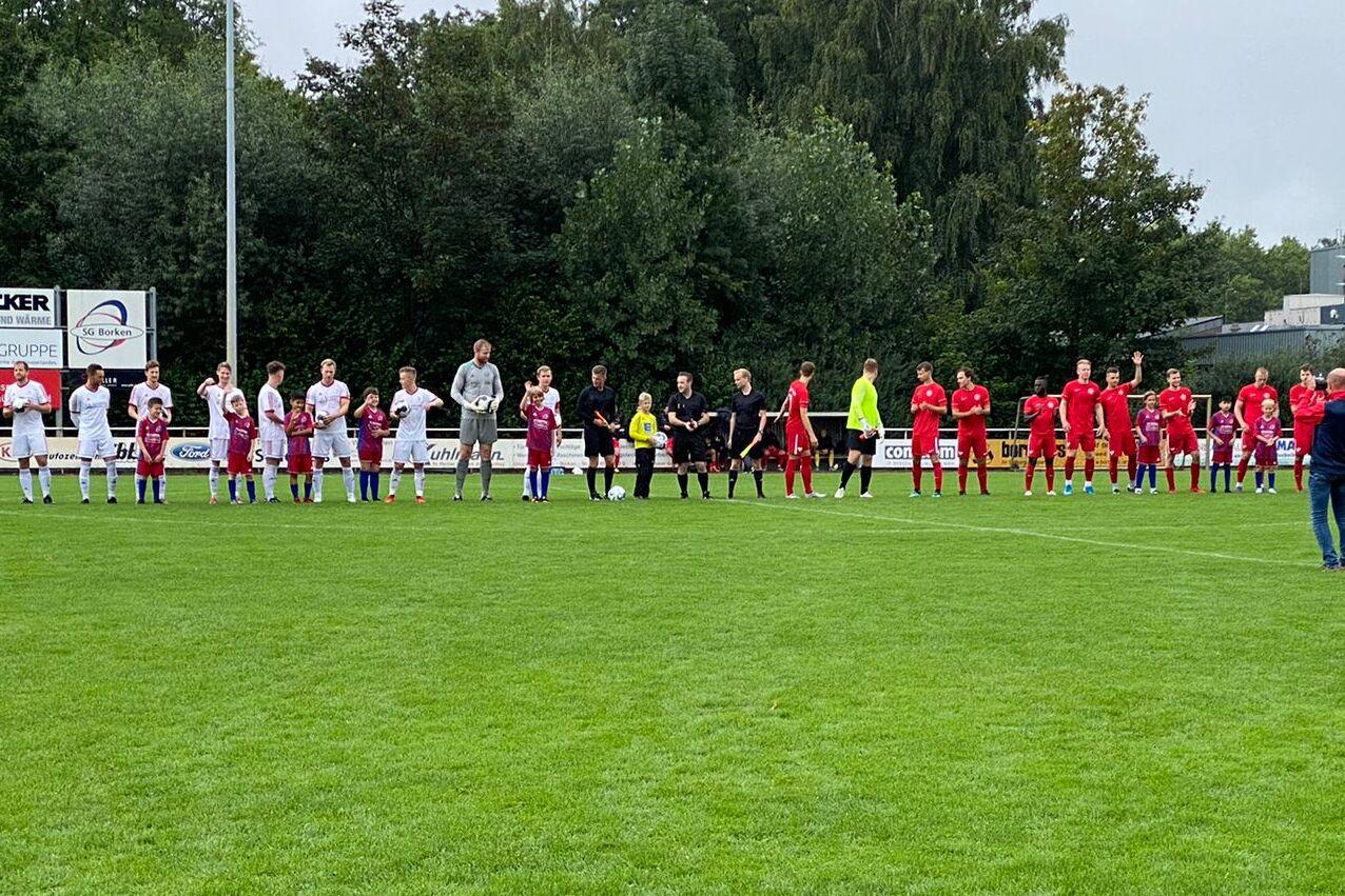 Zwei Fu&szlig;ballmannschaften stehen in einer Reihe auf einem gr&uuml;nen Rasenplatz, begleitet von einigen Zuschauern.