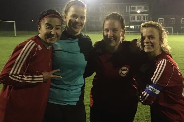 Vier Frauen in Sportkleidung stehen l&auml;chelnd auf einem Fu&szlig;ballfeld bei Nacht, begrenzt von nur schwachem Licht.