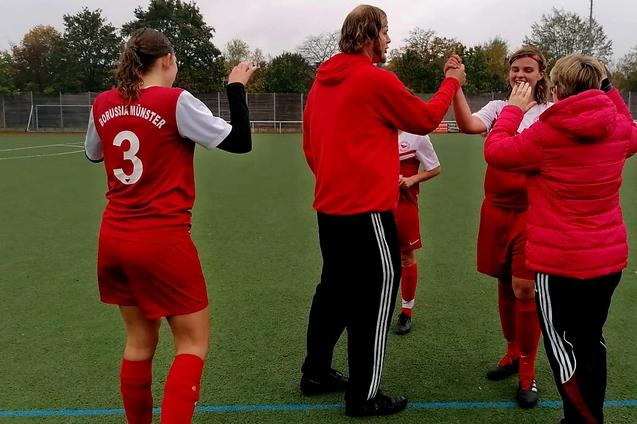 Spielerinnen und Trainer feiern einen Torerfolg auf dem Fu&szlig;ballfeld, umgeben von gr&uuml;ner Rasenfl&auml;che und B&auml;umen im Hintergrund.