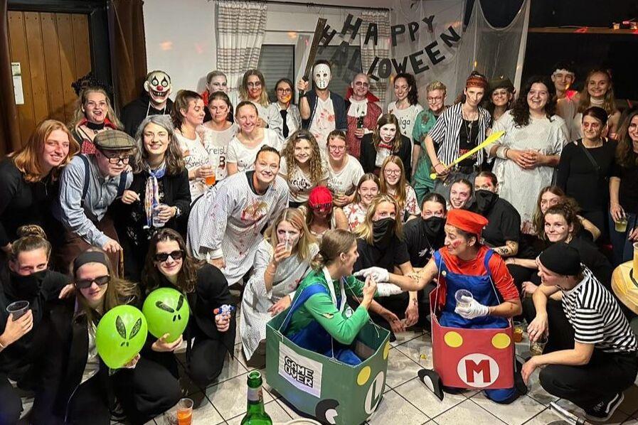 Gruppe von Menschen in Halloween-Kostümen posiert fröhlich vor einer dekorierten Wand mit „Happy Halloween“-Banner.