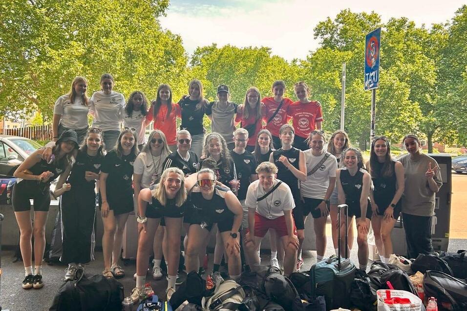 Gruppenfoto von 26 Sportlerinnen in unterschiedlichen Trikots, umgeben von Gepäck, vor einem Baum und Verkehrsschild.