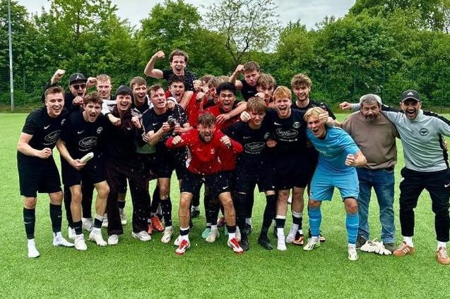 Gruppenshot eines Fu&szlig;ballteams in schwarzen Trikots, jubelnd auf dem Feld, mit einem Pokal in der Mitte.