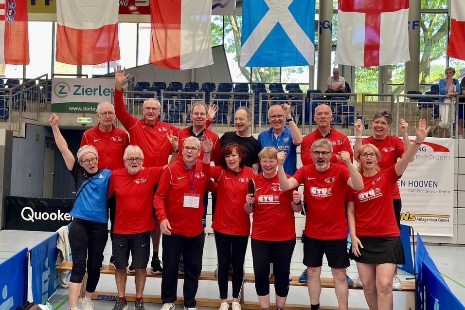 Gruppe von Tischtennisspielern in roten Shirts, die jubelnd in einer Sporthalle stehen, umgeben von Flaggen.