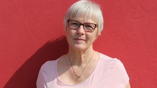Frau mit kurzen, blonden Haaren und Brille steht vor einer roten Wand und l&auml;chelt leicht.