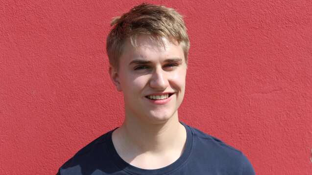 Portr&auml;t eines l&auml;chelnden jungen Mannes vor einer roten Wand, tr&auml;gt ein dunkelblaues T-Shirt mit einem Alien-Logo.