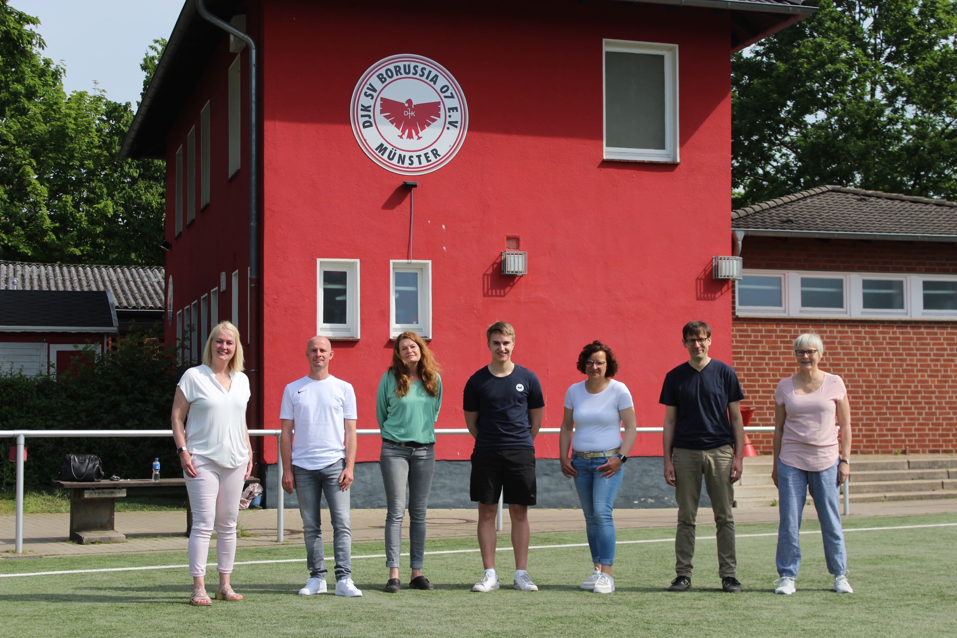 Gruppe von sieben Personen steht auf einem Fu&szlig;ballfeld vor einem roten Vereinsgeb&auml;ude mit Logo.