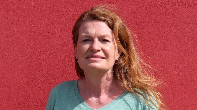 Frau mit langen, lockigen Haaren in einem hellgr&uuml;nen Pullover vor einer roten Wand.