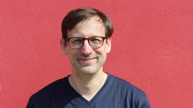 Mann mit Brille und kurzen Haaren steht vor einer roten Wand und l&auml;chelt in die Kamera.