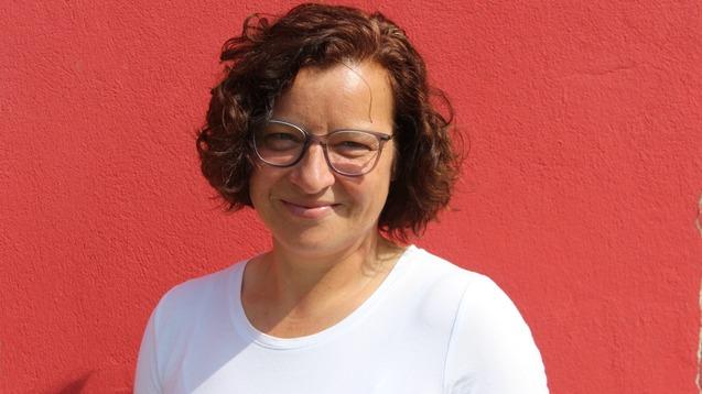 Frau mit lockigem Haar und Brille steht vor einer roten Wand, l&auml;chelt und tr&auml;gt ein wei&szlig;es T-Shirt.