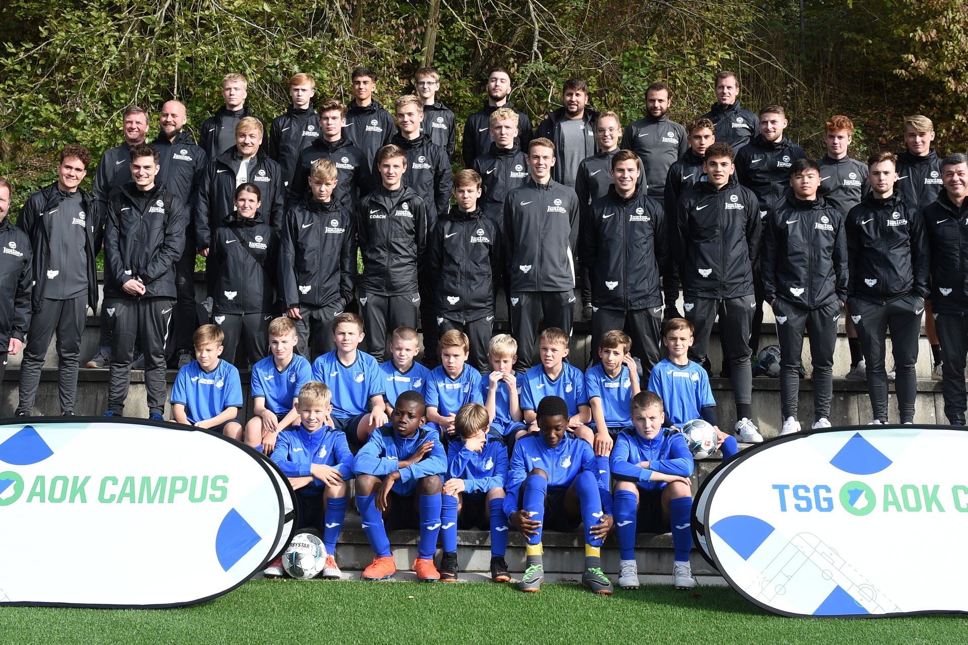 Gruppenfoto einer Fu&szlig;ballmannschaft auf einer Wiese, Trainer und Spieler in schwarzen und blauen Trikots, Banner im Vordergrund.