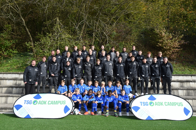 Gruppenfoto einer Fu&szlig;ballmannschaft auf einer Wiese, Trainer und Spieler in schwarzen und blauen Trikots, Banner im Vordergrund.