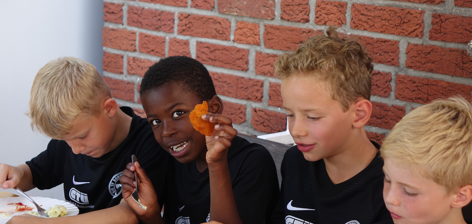 Vier Kinder in schwarzen Trikots sitzen an einem Tisch und essen. Ein Junge h&auml;lt ein St&uuml;ck Gem&uuml;se hoch.