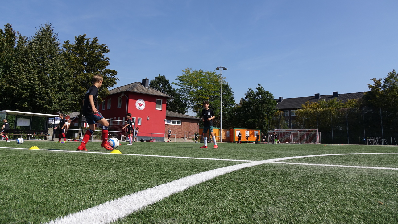 Zwei Kinder &uuml;ben dribbeln mit einem Fu&szlig;ball auf einem Kunstrasenplatz, im Hintergrund ein rotes Geb&auml;ude.