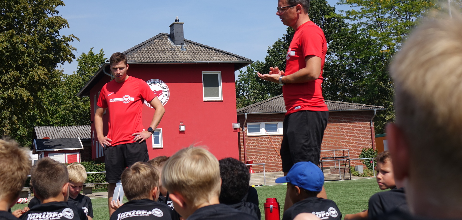 Zwei Trainer in roten T-Shirts geben Kindern auf einem Sportplatz Anweisungen vor einem roten Geb&auml;ude.