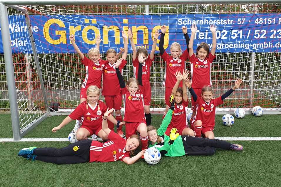 Gruppe von M&auml;dchen in roten Fu&szlig;balltrikots posiert fr&ouml;hlich vor einem Fu&szlig;balltor, umgeben von Fu&szlig;b&auml;llen.