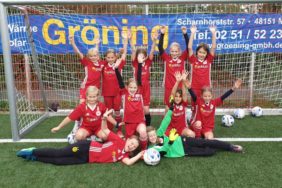 Gruppe von M&auml;dchen in roten Fu&szlig;balltrikots posiert fr&ouml;hlich vor einem Fu&szlig;balltor, umgeben von Fu&szlig;b&auml;llen.
